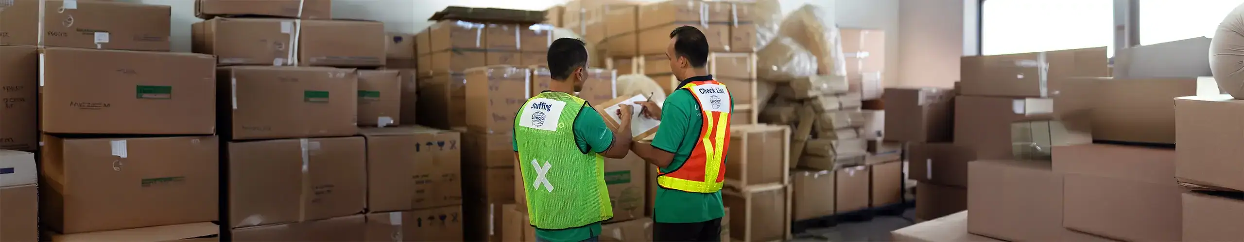 Two workers in safety vests managing and inspecting packages in a warehouse filled with stacked cardboard boxes, showcasing professional cargo logistics services.